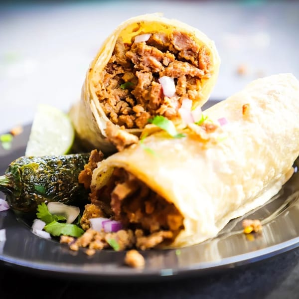 Two burritos filled with meat, onions, and cilantro, served with a grilled jalapeño and lime.