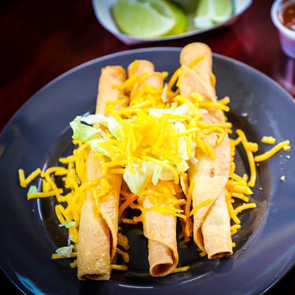 Four crispy taquitos topped with shredded cheese and lettuce on a dark plate, with limes and salsa in the background.