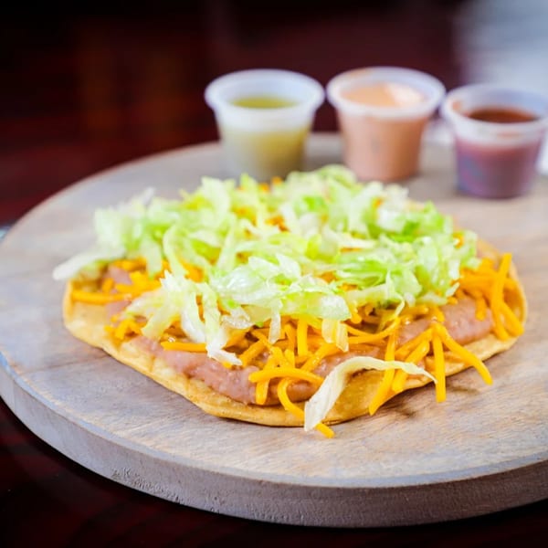 A tostada with refried beans, shredded cheese, and lettuce, served with three dipping sauces.