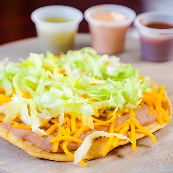 A Taco Plus tostada with refried beans, shredded cheese, and lettuce, with three sauces in the background.